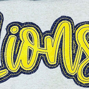May include: A yellow "Lions" word art design with a dark blue outline. The design is on a white fabric background.