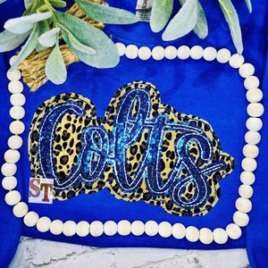 May include: Royal blue long-sleeve shirt with a large "Colts" design. The design features a leopard print background with blue sequin accents and white script lettering. The shirt is decorated with wooden beads and greenery.
