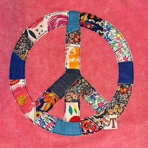 May include: A pink fabric background with a large peace sign made of colorful patchwork fabric. The peace sign is outlined in black thread.