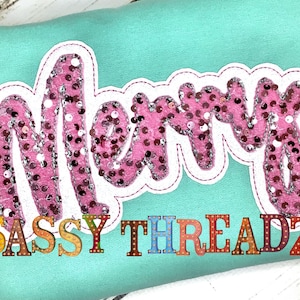 May include: A pink sparkly "Merry" applique with a white outline on a turquoise background. The text "SASSY THREADZ" is below the applique in colorful block letters.