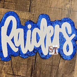 May include: A blue and white sequined patch with the word "Raiders" in white lettering. The patch is made of sequins and has a sparkly finish.