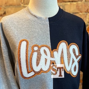 以下が含まれることがあります： グレーとネイビーのトレーナーに、白とオレンジのグリッターで「Lions」のデザインが施されています。