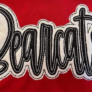 Könnte beinhalten: Roter Stoff mit dem Wort "Bearcats" in schwarzer, handgeschriebener Schrift, umrandet von einem schwarzen Rand. Das Wort besteht aus weißen Pailletten. Ein kleines "ST"-Logo befindet sich in der unteren rechten Ecke.