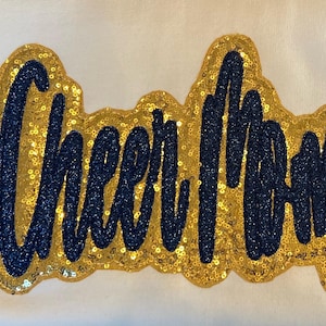 May include: Gold and navy blue sequined patch with the word "Cheer Mom" in a cursive font.