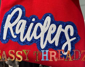May include: Red fabric with a white and blue sequined patch that says "Raiders". Below the patch is a smaller text that says "SASSY THREADZ".