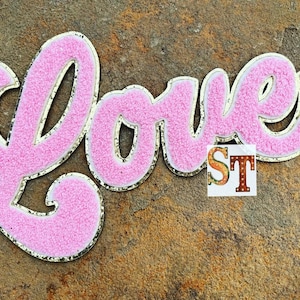 May include: Pink felt "Love" sign with gold glitter trim. The letters are in a cursive font.