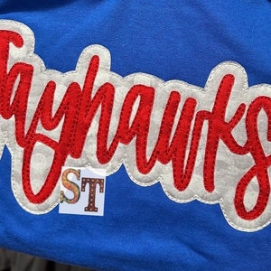 Puede incluir: Parche bordado en rojo y blanco con la palabra "Jayhawks" en letra cursiva. El parche está en una camiseta azul.