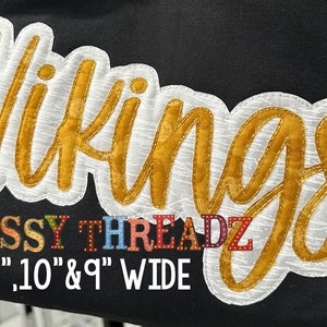 Può includere: Applique dorata "Vikings" con cuciture bianche su uno sfondo di tessuto nero. L'applique è larga 11", 10" e 9". Il testo "SASSY THREADZ" si trova sotto l'applique in lettere multicolori.
