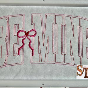 May include: Embroidered design on white fabric with pink thread. The design says "BE MINE" in a large, bold font with a pink bow in the space between the "E" and the "M".