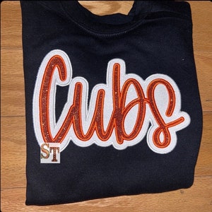 May include: A black sweatshirt with a white and orange glitter iron-on patch that says "CUBS".