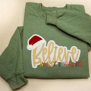 May include: Green sweatshirt with a gold "Believe" design and a red Santa hat applique. The text "Sassy Threadz" is below the design.