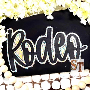 May include: Black fabric with a glittery iridescent "Rodeo" applique. The applique is outlined in black thread.
