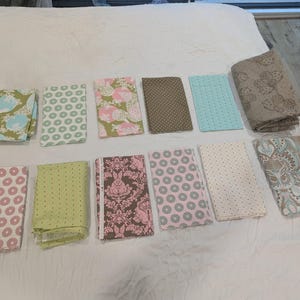 May include: A collection of twelve rectangular fabric swatches arranged in two rows. The patterns include floral, geometric, and damask designs in various colours such as pink, green, brown, blue, and cream. Each piece of fabric is folded flat.