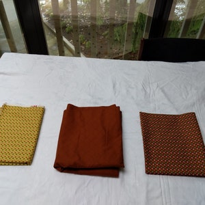 Op de afbeelding: Drie gevouwen stukken stof naast elkaar geplaatst. Het linker stuk is geel met een klein rood en groen geometrisch patroon. Het middelste stuk is effen donker roestbruin. Het rechter stuk is donkerbruin met een klein rood en groen patroon.