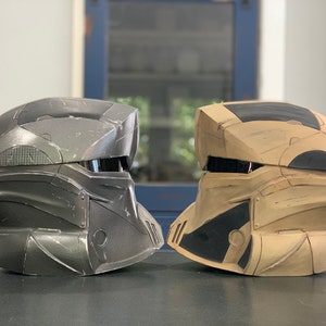 Puede incluir: Dos cascos de estilo futurista, uno gris oscuro y otro beige con detalles negros, se muestran uno al lado del otro. Los cascos tienen un &aacute;rea de visera y paneles detallados, lo que sugiere un tema de ciencia ficci&oacute;n o cosplay.