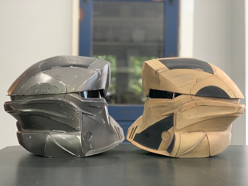 Puede incluir: Dos cascos de estilo futurista, uno gris oscuro y otro beige con detalles negros, se muestran uno al lado del otro. Los cascos tienen un &aacute;rea de visera y paneles detallados, lo que sugiere un tema de ciencia ficci&oacute;n o cosplay.