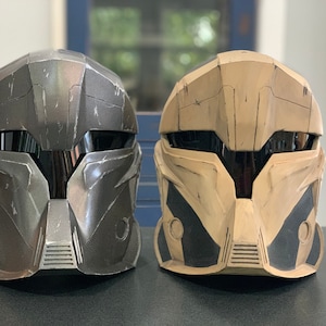 Puede incluir: Se muestran dos cascos futuristas, uno gris oscuro y otro beige, uno al lado del otro. Los cascos tienen un &aacute;rea de visera y dise&ntilde;os angulares. El casco gris oscuro tiene una apariencia texturizada, mientras que el casco beige tiene detalles en negro.