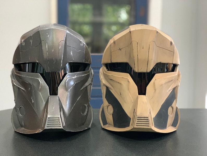 Puede incluir: Se muestran dos cascos futuristas, uno gris oscuro y otro beige, uno al lado del otro. Los cascos tienen un &aacute;rea de visera y dise&ntilde;os angulares. El casco gris oscuro tiene una apariencia texturizada, mientras que el casco beige tiene detalles en negro.