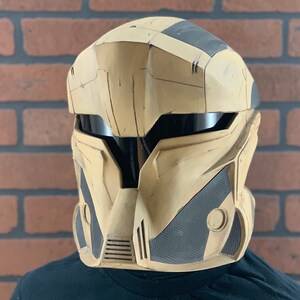 Puede incluir: Un casco futurista beige y negro con visera. El casco tiene un dise&ntilde;o segmentado con elementos angulares y un panel de malla en la parte inferior delantera. Probablemente sea para cosplay o disfraces.