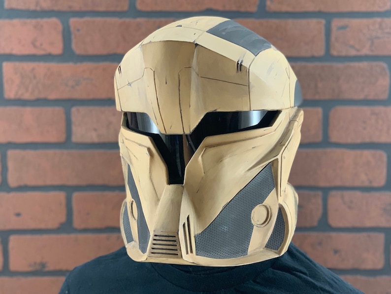 Puede incluir: Un casco futurista beige y negro con visera. El casco tiene un dise&ntilde;o segmentado con elementos angulares y un panel de malla en la parte inferior delantera. Probablemente sea para cosplay o disfraces.