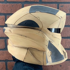 Puede incluir: Un casco futurista beige y gris con un dise&ntilde;o segmentado. El casco presenta una visera oscura y paneles texturizados. La est&eacute;tica general sugiere un tema de ciencia ficci&oacute;n o cosplay.