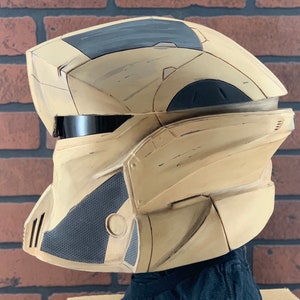 Puede incluir: Un casco futurista beige y gris con una visera negra. El casco tiene un dise&ntilde;o geom&eacute;trico con paneles angulares y una secci&oacute;n gris texturizada en el lateral. El casco es probablemente para cosplay o disfraces.
