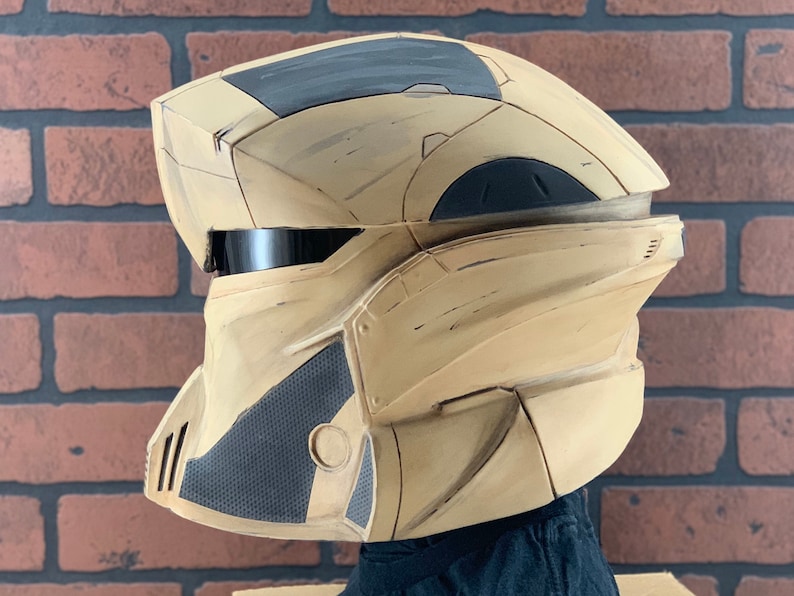 Puede incluir: Un casco futurista beige y gris con una visera negra. El casco tiene un dise&ntilde;o geom&eacute;trico con paneles angulares y una secci&oacute;n gris texturizada en el lateral. El casco es probablemente para cosplay o disfraces.