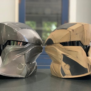 Puede incluir: Dos cascos futuristas, uno gris met&aacute;lico y otro beige, se muestran uno al lado del otro. Los cascos tienen una visera y un dise&ntilde;o angular detallado, lo que sugiere un tema de ciencia ficci&oacute;n o cosplay.