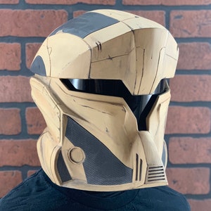 Puede incluir: Un casco futurista en beige y negro con visera. El casco tiene un dise&ntilde;o segmentado con un aspecto texturizado. La visera es oscura y el casco parece estar hecho de un material duradero. El casco est&aacute; dise&ntilde;ado para ser usado.