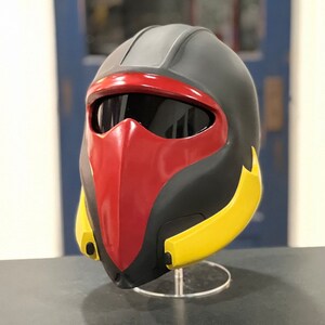 Puede incluir: Un casco negro, rojo y amarillo con visera. El casco tiene un diseño elegante con una máscara facial roja, cuerpo negro y detalles amarillos. La visera es oscura y el casco se exhibe sobre un soporte transparente.