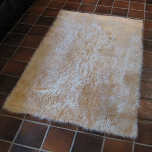 May include: A fluffy, cream-colored rug with a soft, plush texture. The rug is laid out on a tiled floor.