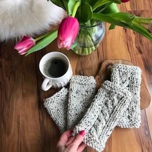 Könnte beinhalten: Drei Paar gestrickte Fingerlose Handschuhe in hellgrau mit einem Zopfmuster. Die Handschuhe liegen auf einer Holzoberfläche mit einer Tasse Kaffee und rosa Blumen im Hintergrund.