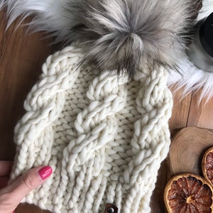 Op de afbeelding: Witte gebreide beanie met een grote, pluche pompon en een bruin leren label met de tekst "Rosehip Lane".