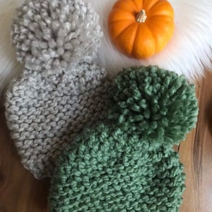 K&ouml;nnte beinhalten: Zwei Strickm&uuml;tzen mit flauschigen Bommeln. Eine M&uuml;tze ist grau und die andere gr&uuml;n.