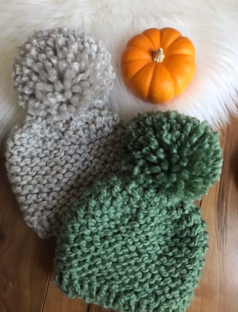 K&ouml;nnte beinhalten: Zwei Strickm&uuml;tzen mit flauschigen Bommeln. Eine M&uuml;tze ist grau und die andere gr&uuml;n.