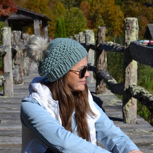 Könnte beinhalten: Eine Frau trägt eine türkisfarbene Strickmütze mit einem weißen Bommel oben. Sie sitzt auf einer Holzbrücke mit einem Holzgeländer. Der Hintergrund ist ein verschwommenes Bild von Bäumen und einer Holzkonstruktion.