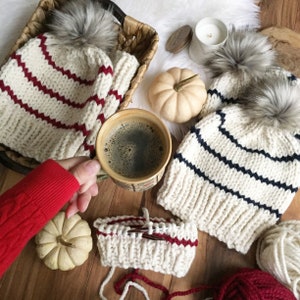 May include: Two white and navy striped knit hats with pom poms, one white and red striped knit hat, and a cup of coffee on a wooden surface. There is also a white pumpkin and yarn balls in the image.