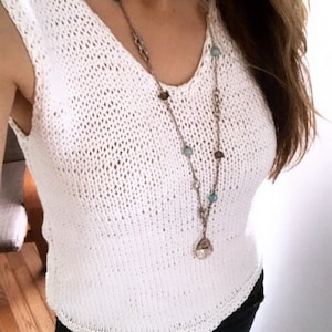 May include: A white, sleeveless, V-neck knit top. A long silver necklace with blue and brown beads and a teardrop pendant hangs over the top. The top is paired with dark-colored pants.