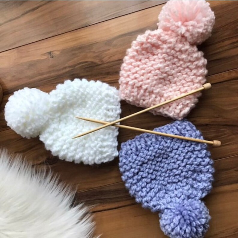 K&ouml;nnte beinhalten: Drei gestrickte M&uuml;tzen mit Bommeln in Wei&szlig;, Rosa und Lila. Jede M&uuml;tze ist mit einer Stricknadel aus Holz kombiniert.