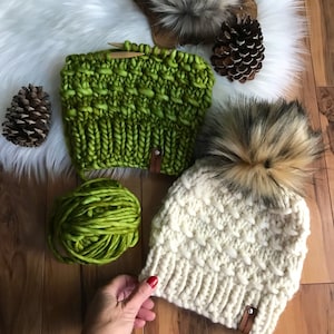 May include: Two knitted hats, one green and one white with a faux fur pom pom. The green hat is made with a chunky yarn and has a simple stitch pattern. The white hat has a more intricate stitch pattern and is made with a finer yarn. Both hats have leather labels.
