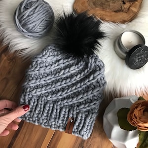 May include: Gray knitted beanie with a black faux fur pom pom. The beanie has a brown leather tag with the text "Knit Love" on it.