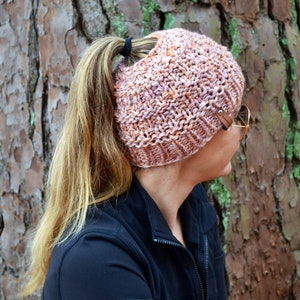 Può includere: Un cappello di lana rosa e arancione con un foro per la coda di cavallo. Il cappello ha un cinturino in pelle marrone con una fibbia in metallo.