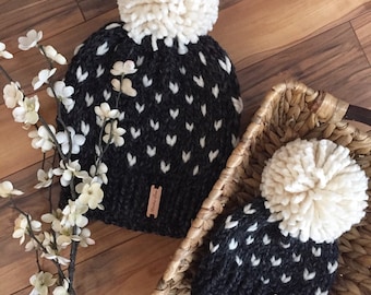 Könnte beinhalten: Zwei schwarze und weiße Strickmützen mit weißen Bommeln. Die Mützen haben ein weißes Herzmuster und ein braunes Leder-Etikett mit dem Text "Handmade Love" darauf.