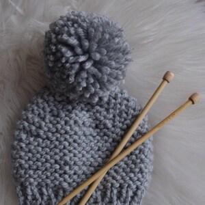 K&ouml;nnte beinhalten: Eine graue Strickm&uuml;tze mit einem flauschigen Bommel oben. Zwei Stricknadeln aus Holz liegen auf der M&uuml;tze.