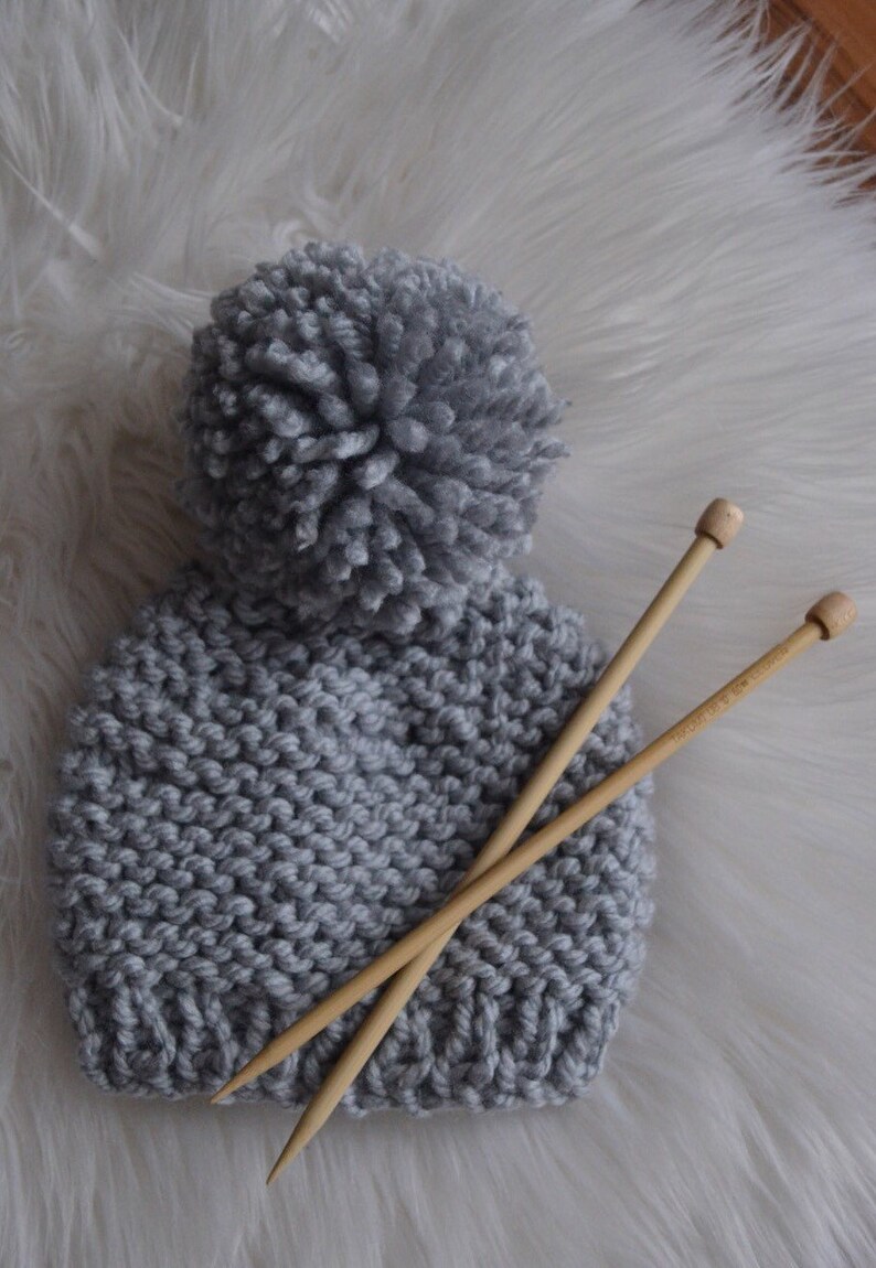 K&ouml;nnte beinhalten: Eine graue Strickm&uuml;tze mit einem flauschigen Bommel oben. Zwei Stricknadeln aus Holz liegen auf der M&uuml;tze.