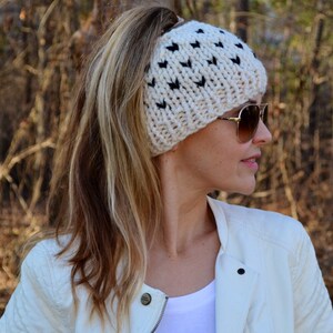Könnte beinhalten: Eine weiße gestrickte Pferdeschwanz-Mütze mit einem schwarzen Herzmuster. Die Mütze wird von einer Person mit langen blonden Haaren und Sonnenbrille getragen.