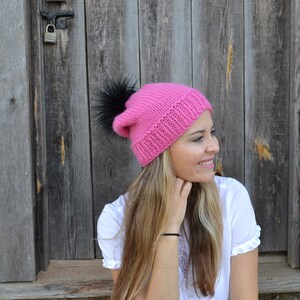 May include: A vibrant pink knitted beanie hat with a black faux fur pom-pom. The hat features a ribbed design and a folded-over brim. The background is a weathered wooden door.