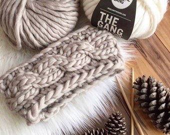 Könnte beinhalten: Nahaufnahme eines gestrickten Stirnbands mit Zopfmuster. Der Stirnband ist aus einem dicken Garn in hellbrauner Farbe gefertigt. Im Hintergrund sind zwei Garnrollen zu sehen, eine weiße und eine hellbraune. Die Garnrollen tragen Etiketten mit der Aufschrift "Wool and the Gang" und "The Gang Collection".