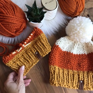 Op de afbeelding: Twee gebreide mutsen in oranje, wit en mosterdgeel. De grotere muts heeft een witte pompon bovenop. Beide mutsen hebben een bruin leren label met de tekst "Pretty Lab" erop.