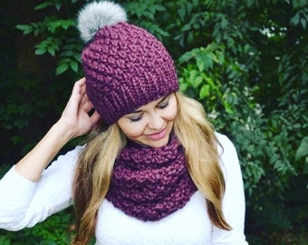 May include: A purple knitted hat with a faux fur pom pom and a matching purple knitted scarf.